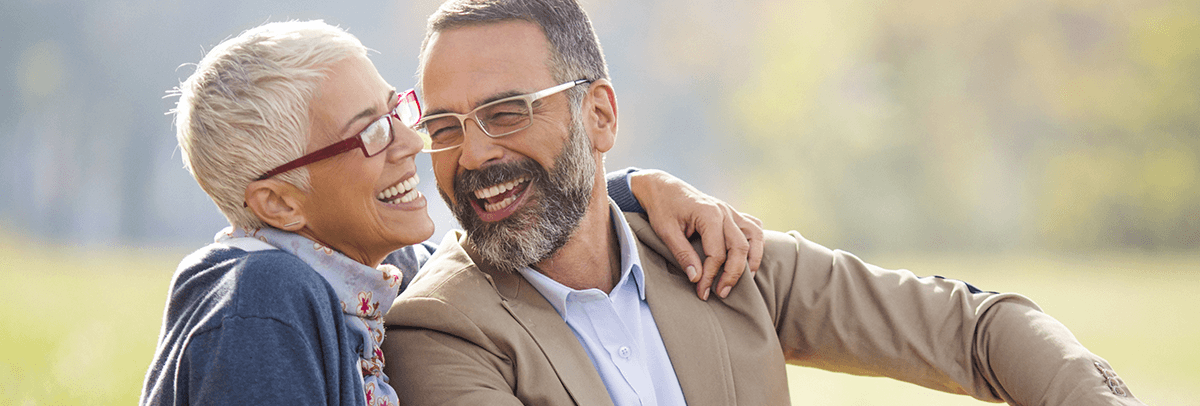 Laughing-Senior-couple-wearing-eyeglasses Smiling iddle-aged couple sitting togehter on grassy hill.